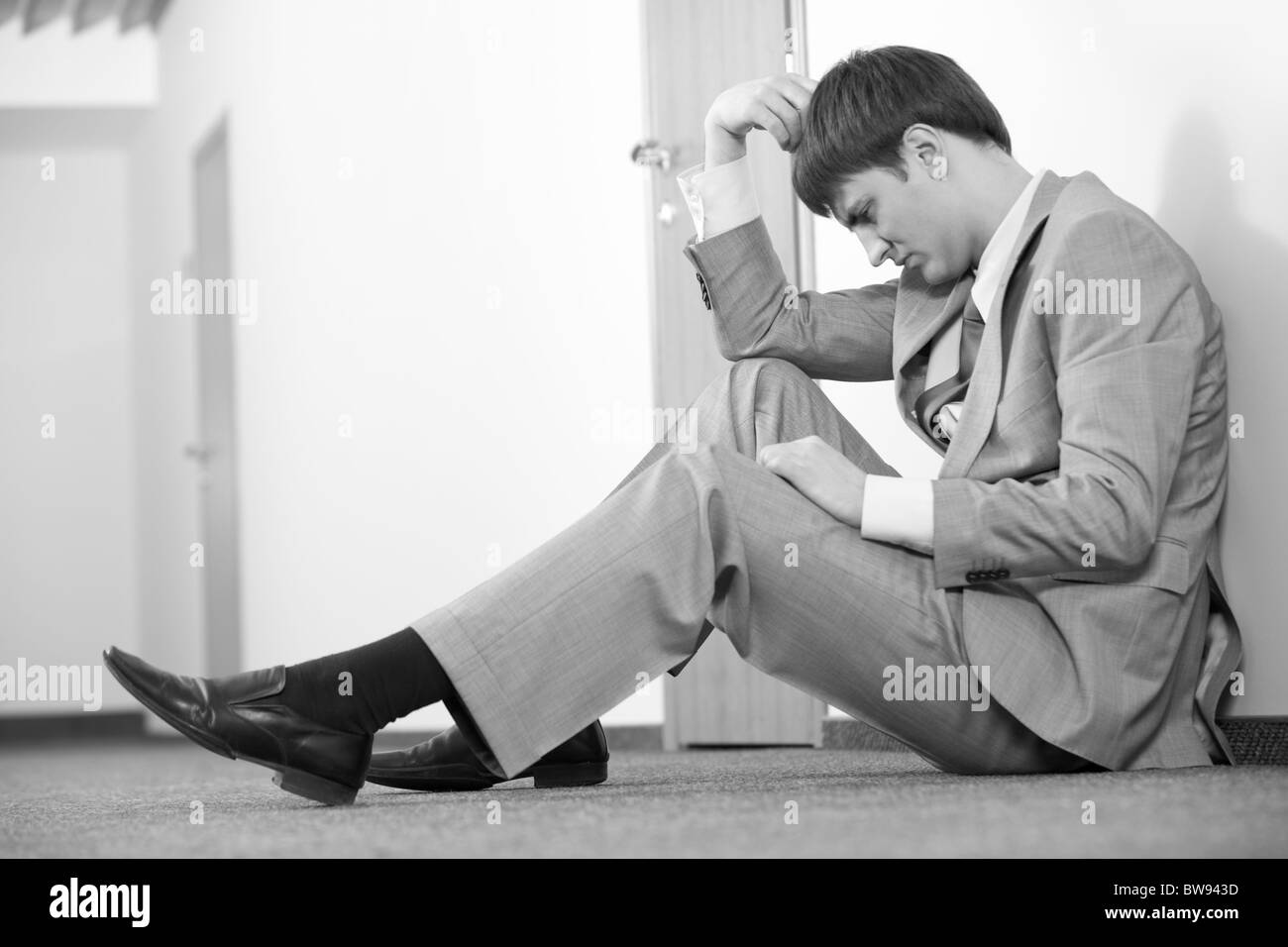 Black and white photo of thinking man sitting on the floor against wall Stock Photo