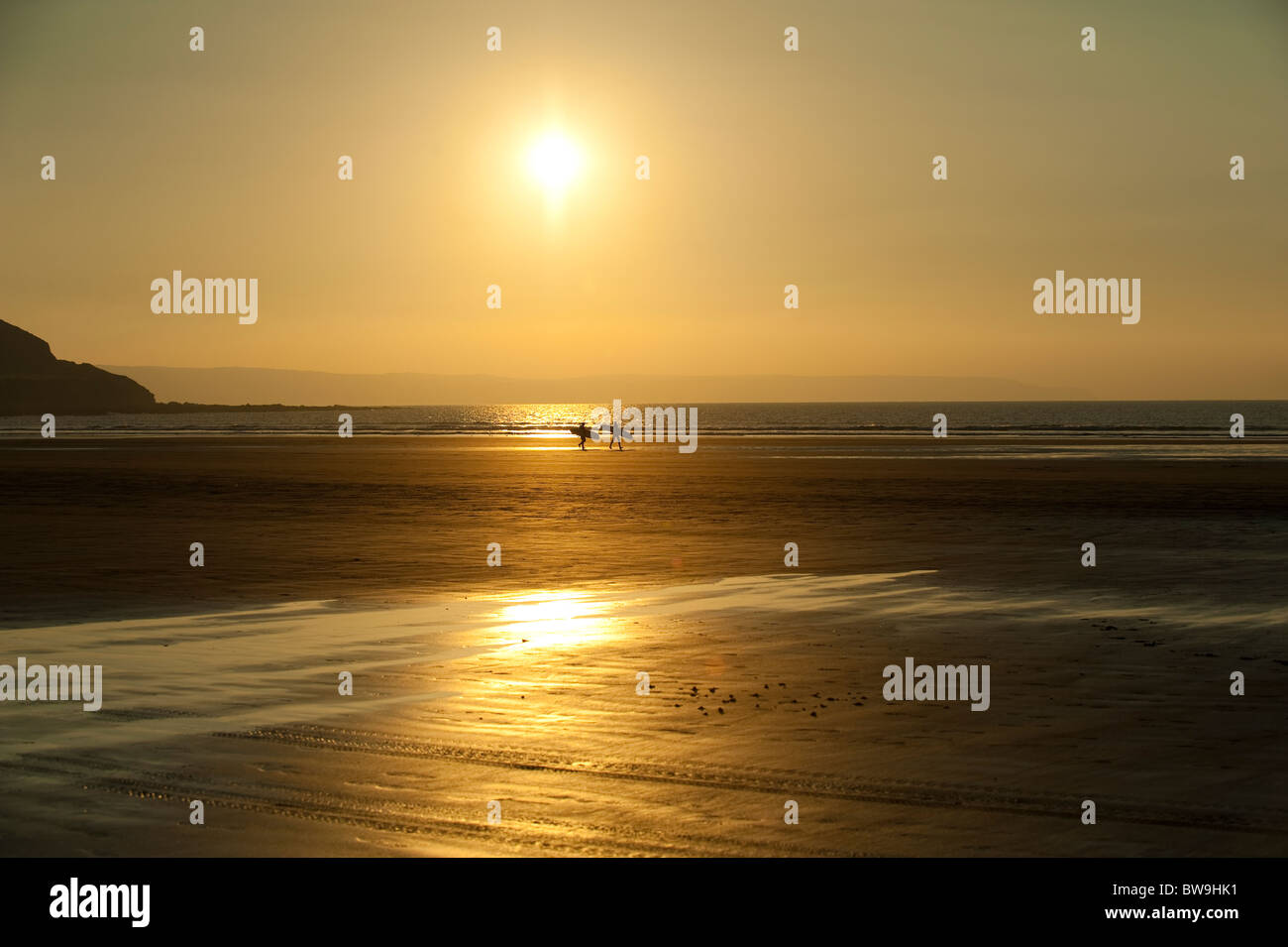 Two surfers carry surf boards into the sunset on beach at Westward Ho! Devon UK Stock Photo