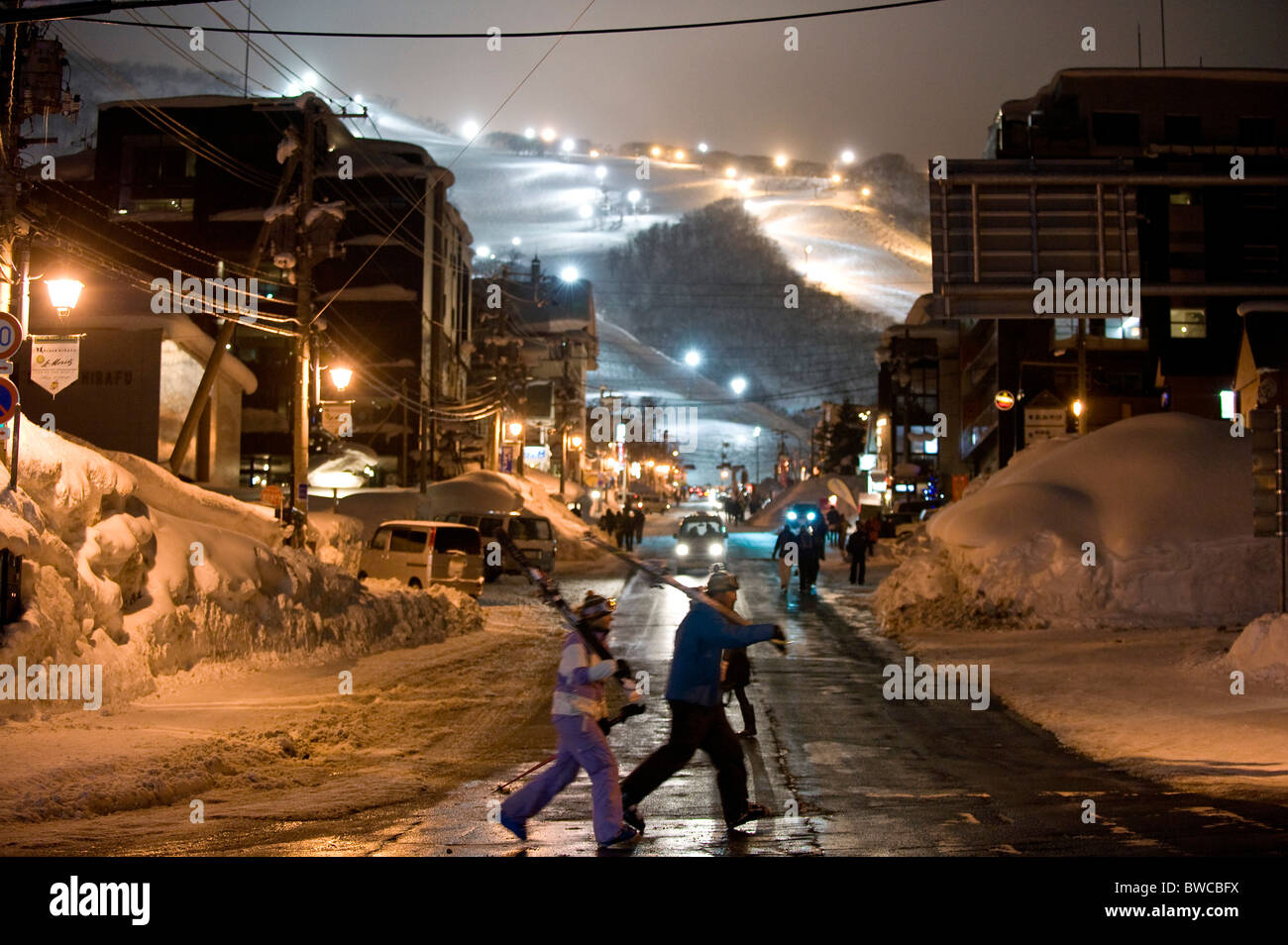The lights of Grand Hirafu ski resort in the distance, boarders and skiers cross the main street at Niseko, Japan Stock Photo