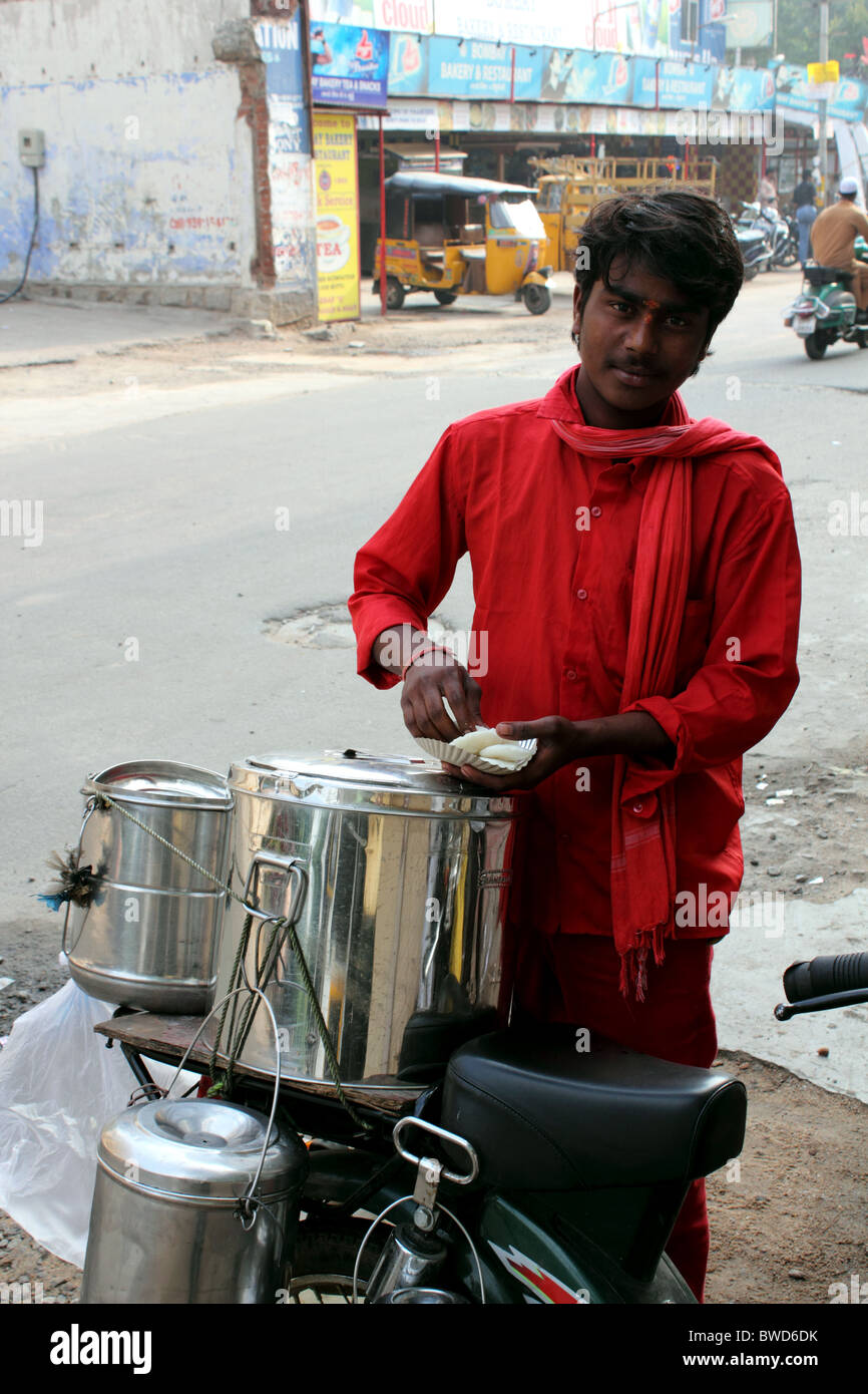 Indian man in red shirt selling food from a bicycle; serving breakfast items from metal containers on a bike pannier Stock Photo