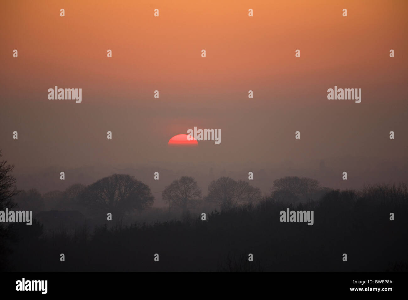 Mysterious floating half sun setting in winter rising mist and fog over a dark wooded valley under red sky near Hawkhurst Kent Stock Photo