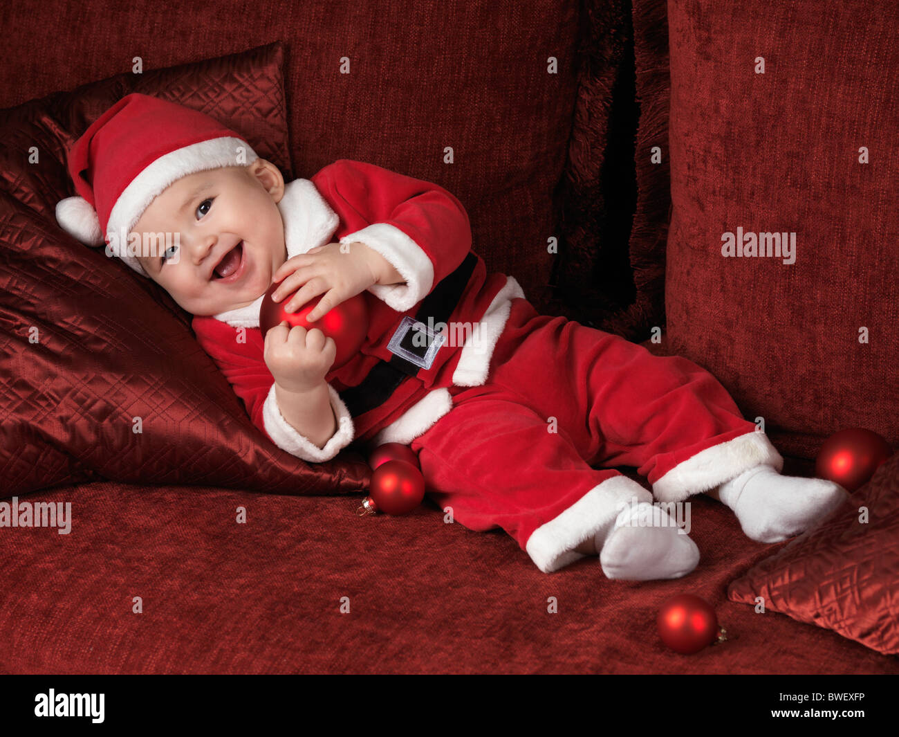 Happy smiling six month old baby boy in Santa Christmas costume lying on a sofa with a red