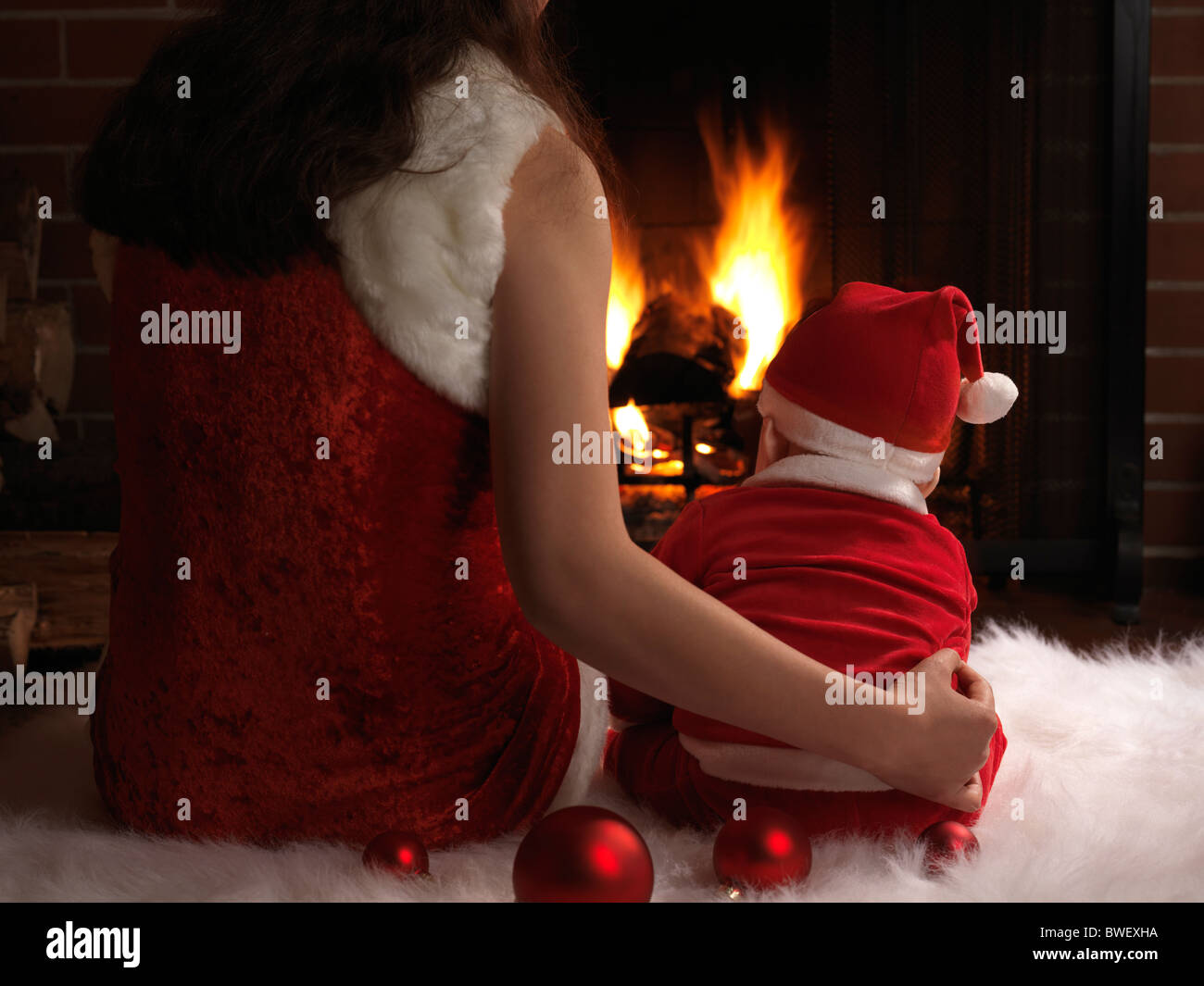 Mother and a little baby boy wearing Santa costumes, sitting in front of a fireplace on Christmas Eve. Stock Photo