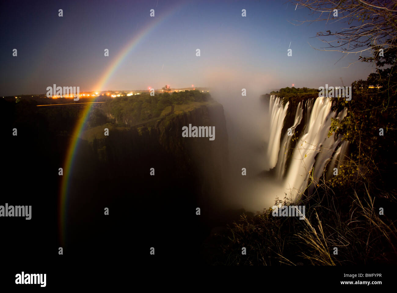 Night scene moon rainbow over deep waterfall on right town lights