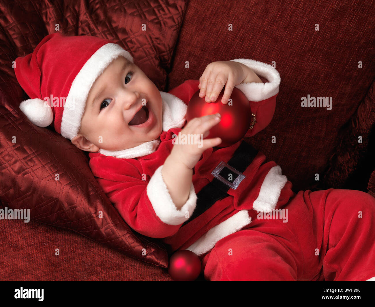 Happy smiling six month old baby boy in Santa Christmas costume lying on a sofa with a red bauble in his hands Stock Photo