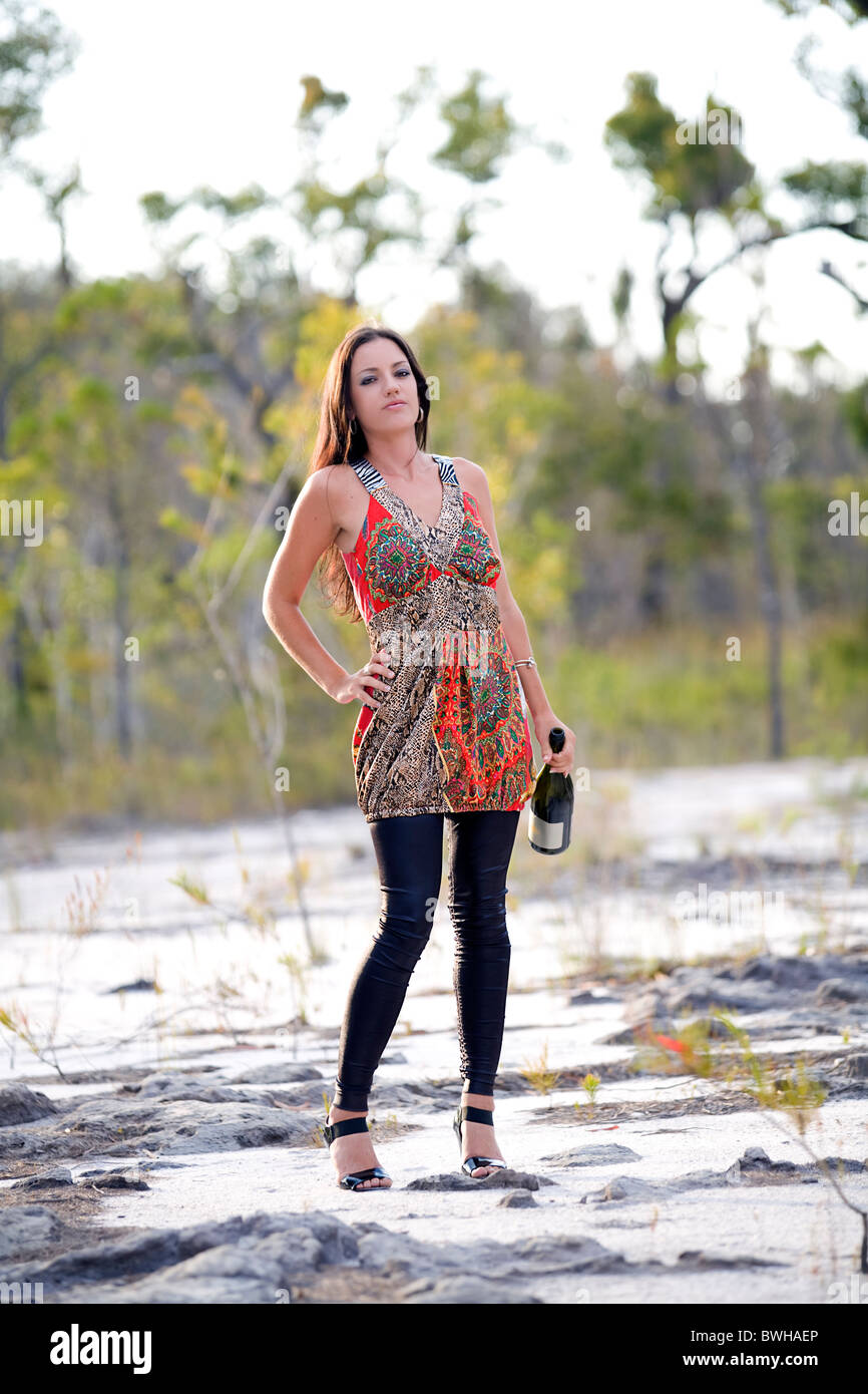 Attractive young brunette woman wearing bright color mini dress stands holding wine bottle in the Australian outback country Stock Photo