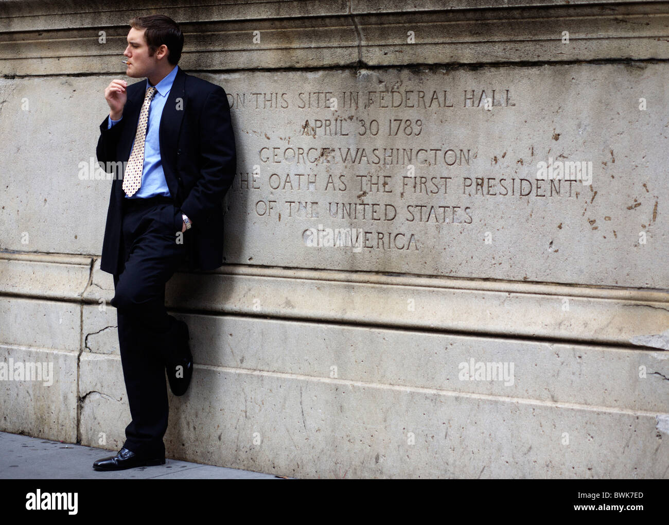 A man in a suit smokes a cigarette outside the Federal Hall on Wall Street in New York City , United States of America Stock Photo
