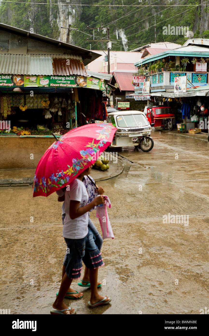 Filipino girls walk through the streets of El Nido under an umbrella during a rain shower. Stock Photo