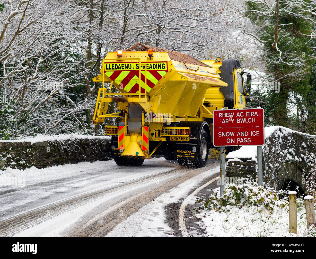 Snow and Ice on Pass road sign, English/Welsh, set in a background of snow covered trees and a gritting snowplough Stock Photo