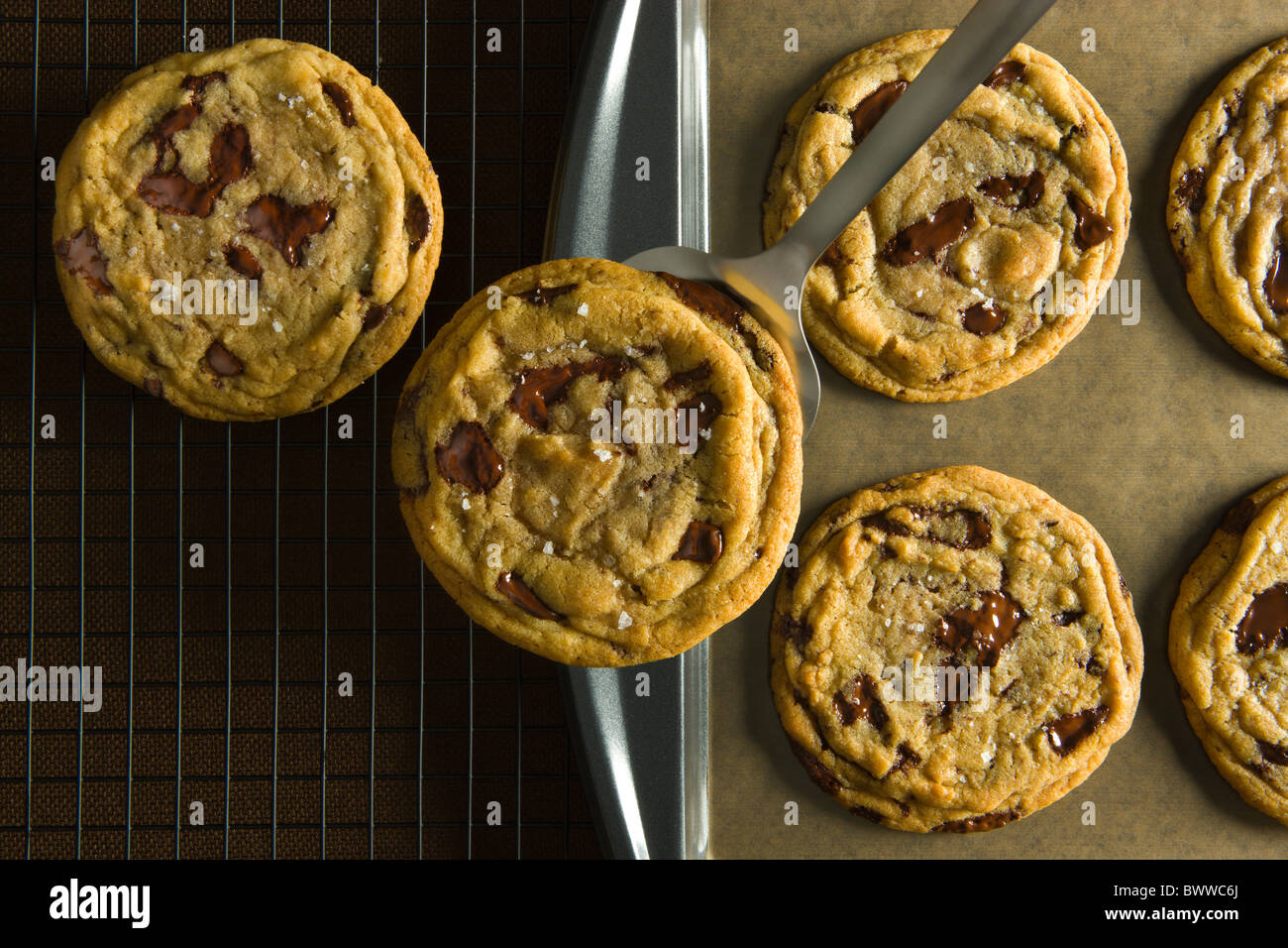 A Spatula removing a Chocolate Chip Cookies from a baking sheet over a brown linen table cloth and a wire cooling rack. Stock Photo