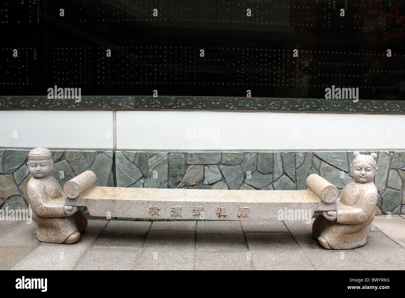 Chinese stone bench supported by carved human figures of boy and girl; behind is black stone with Chinese characters or writing Stock Photo