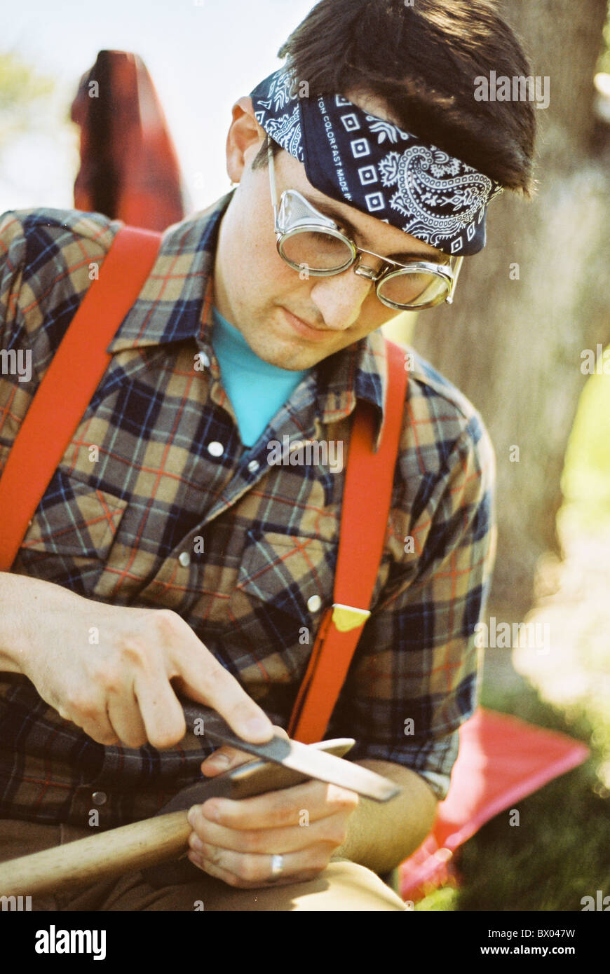 A man wearing suspenders, flannel, and a bandana sharpens a maul. Stock Photo