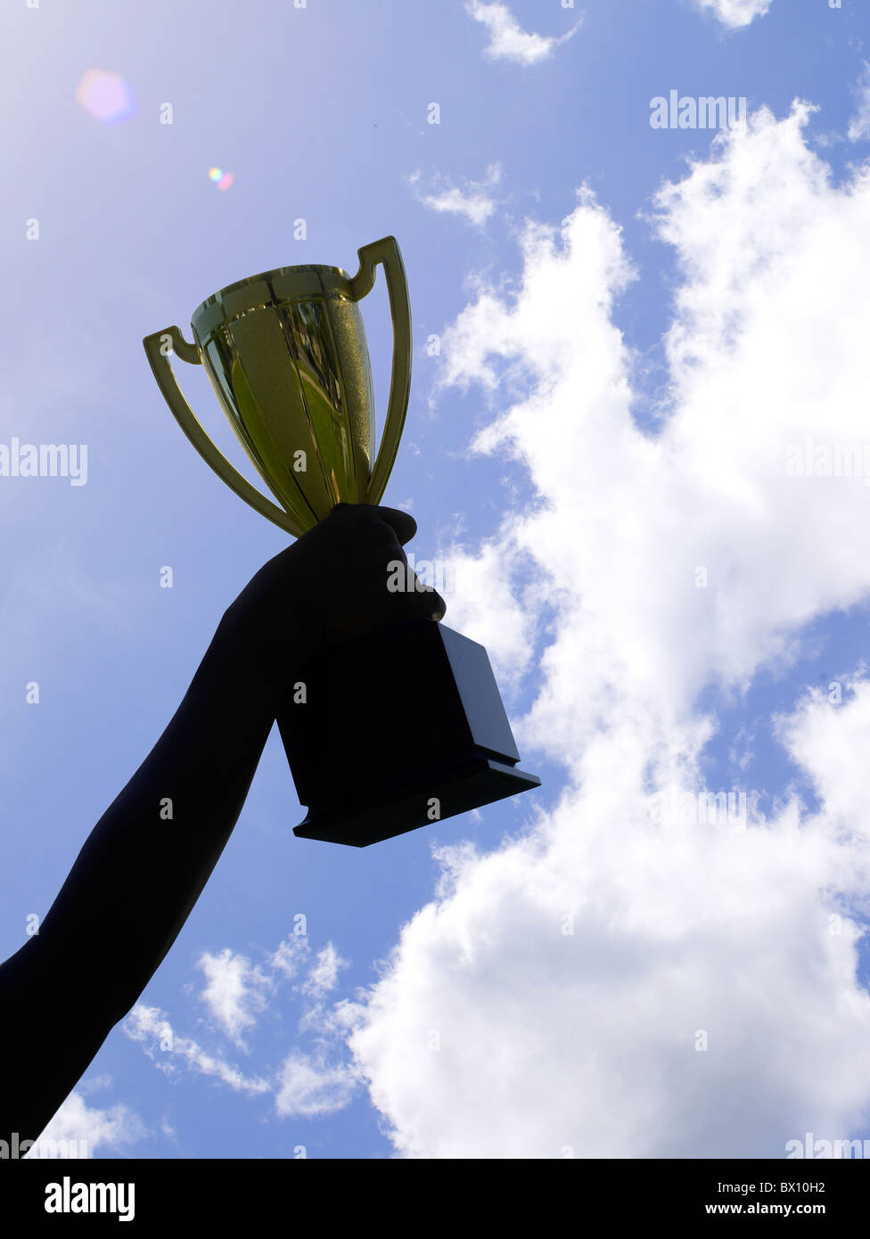 A victorious person holds up a large, gleaming trophy cup, silhouetted against a vivid blue sky with a few wispy clouds. Stock Photo
