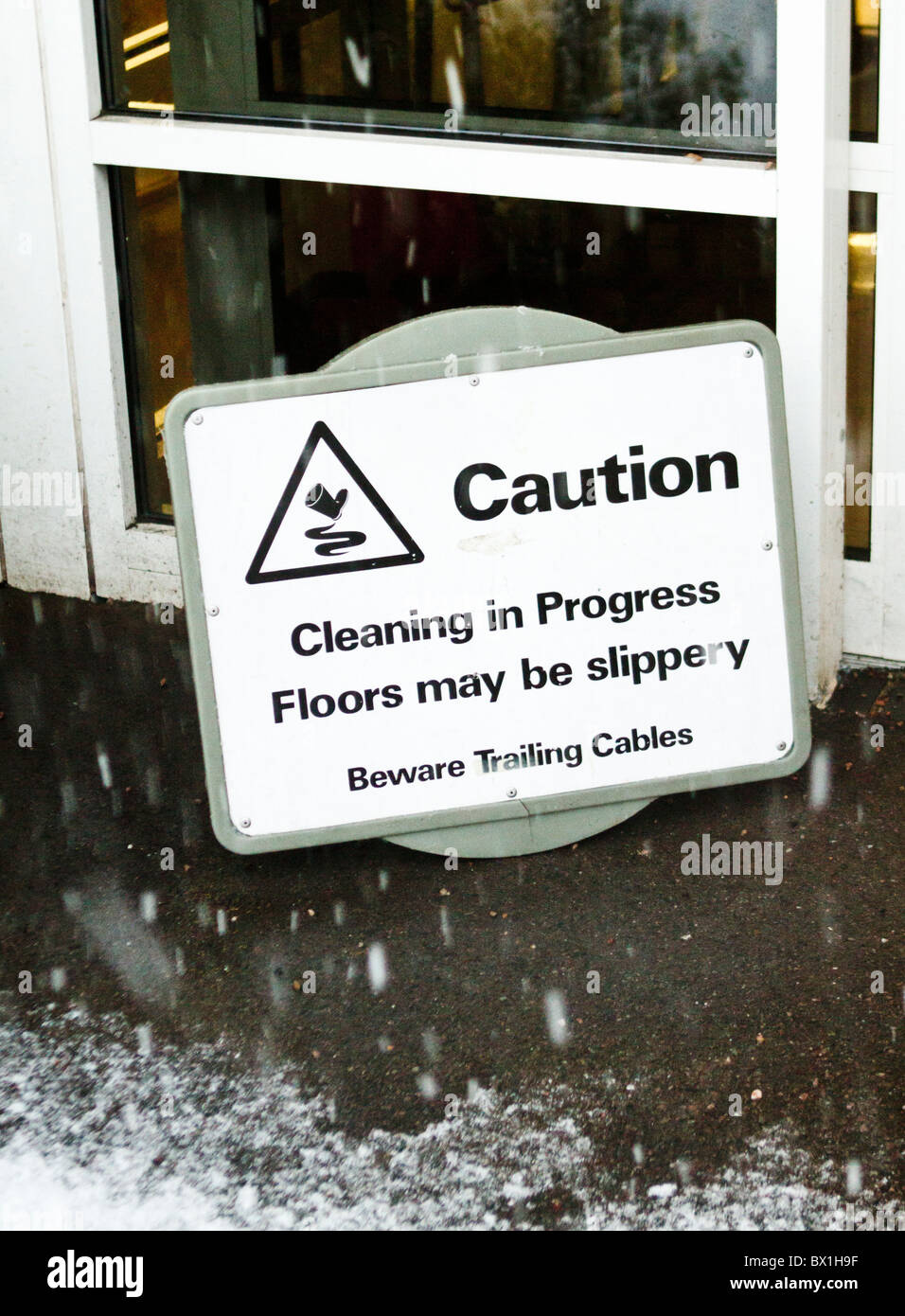 Snow falling around a sign warning of slippery floors due to cleaning