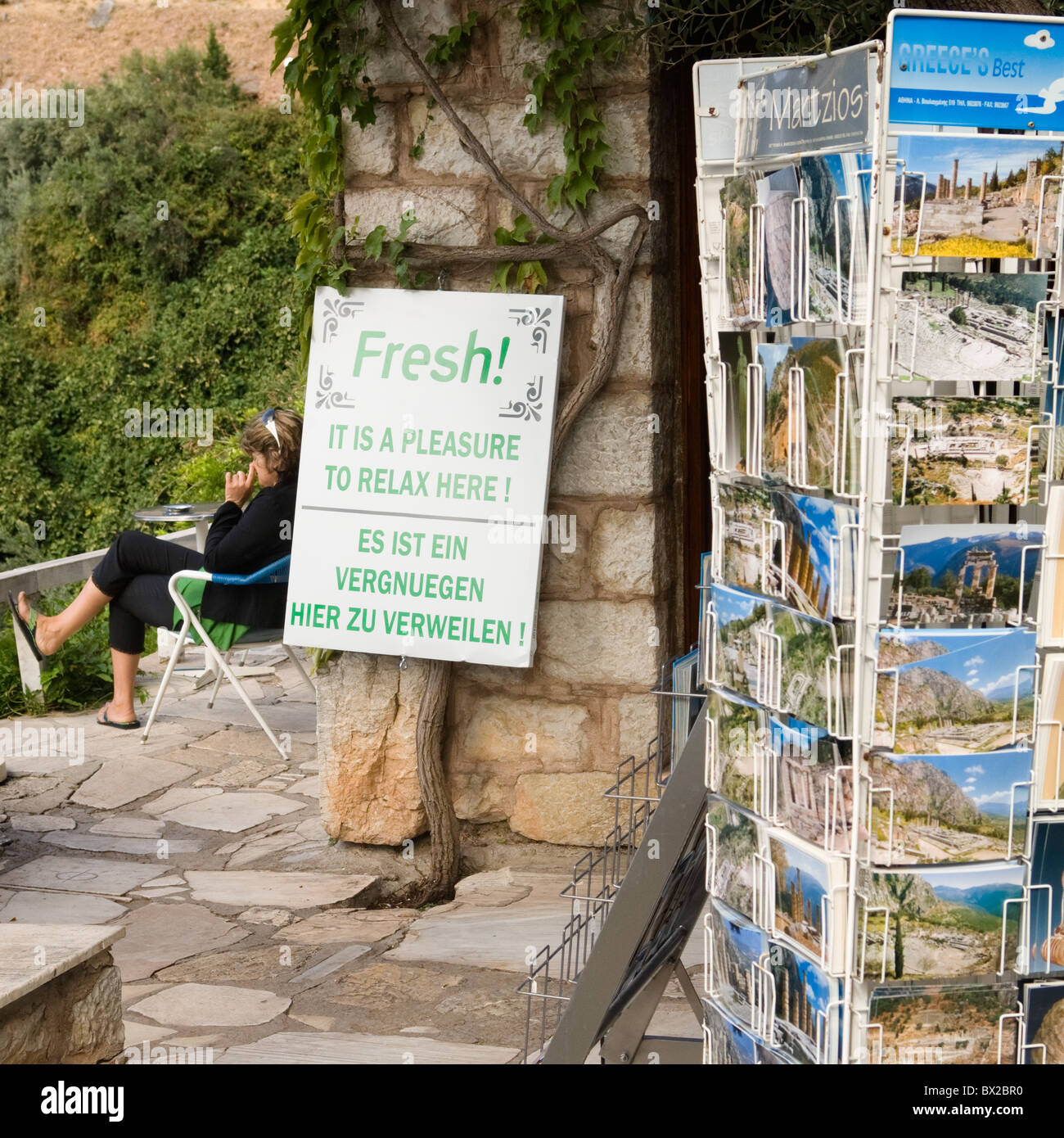 A woman picking her nose next to a sign saying 'Fresh! - It is a pleassure to relax here!' at a cafe in Delphi Greece. Stock Photo