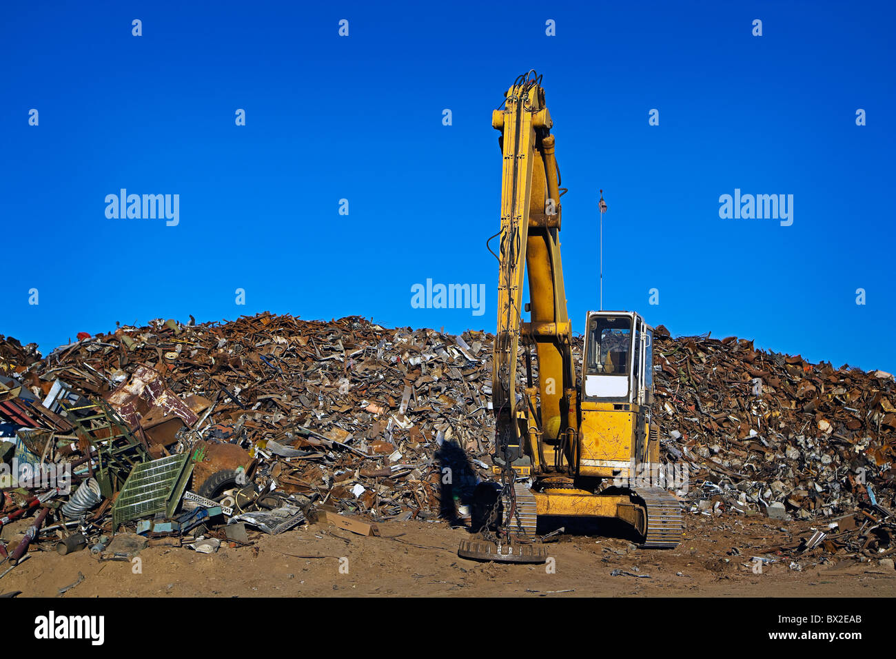 A large yellow crane works with a huge pile of scrap metal, getting it ready for recycling under clear blue skies. Stock Photo