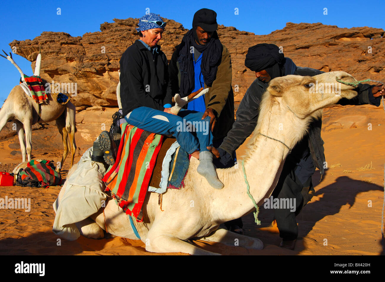 Tuareg nomads helping a female tourist to climb on a dromedary in the Acacous Mountains, Sahara desert, Libya Stock Photo