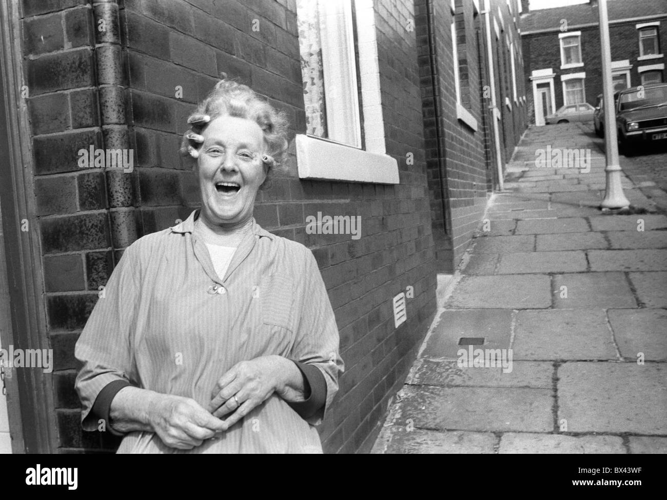 Working class 1980s UK,  white woman in hair curlers wearing her housecoat standing outside her terraced house laughing Blackburn Lancashire England 1983. HOMER SYKES Stock Photo