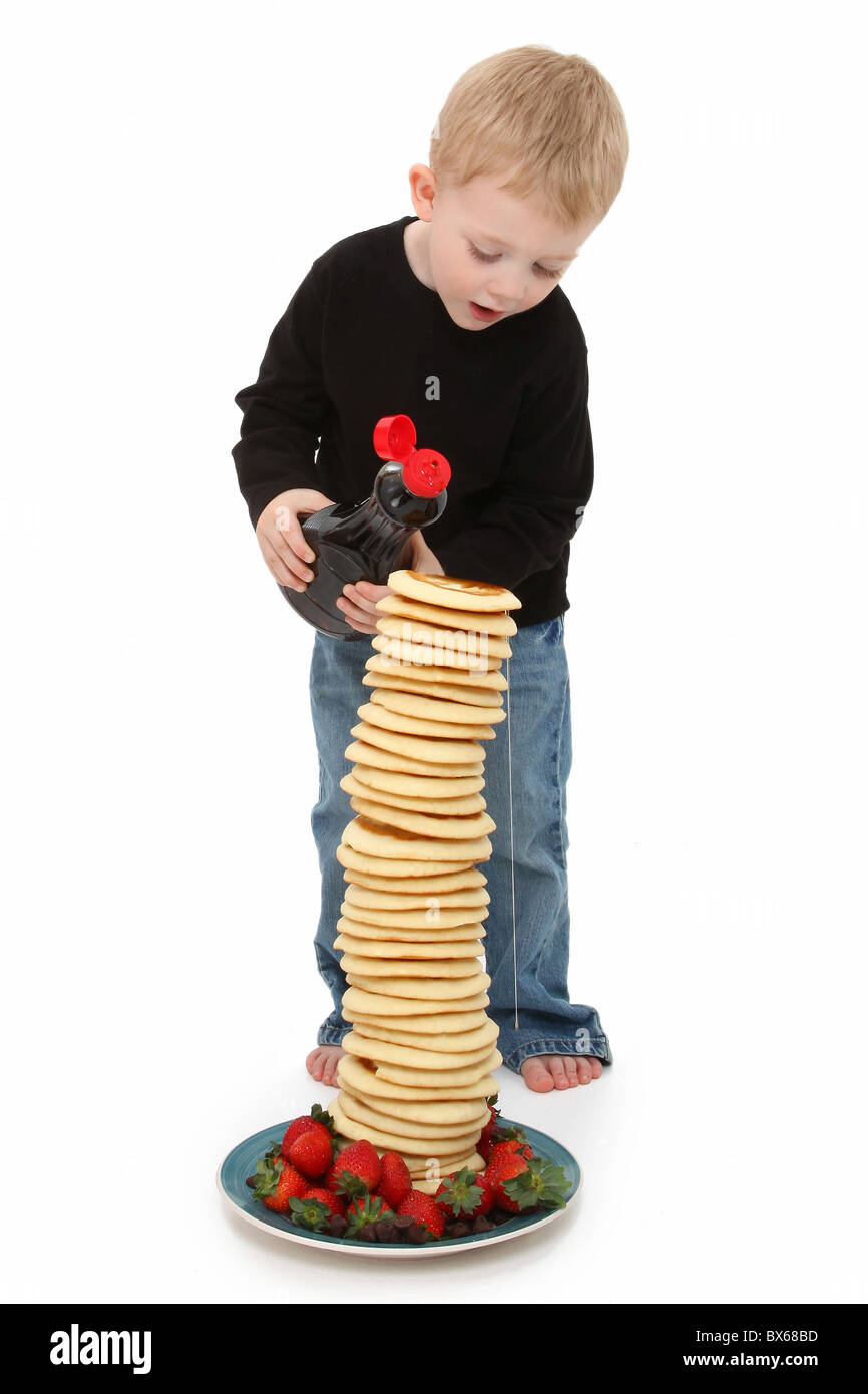 Adorable 2 year old boy pouring syrup on a giant stack of pancakes with strawberries and chocolate over white. Stock Photo
