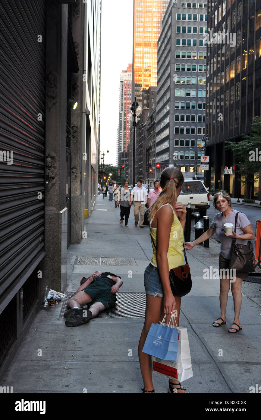 A homeless man lies across a street in New York while two woman tourists stop to show concern for his welfare Stock Photo