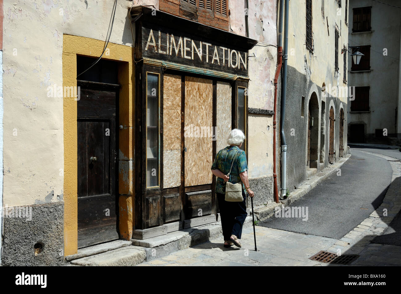 Old Woman with Walking Stick Walks Past a Boarded-Up & Closed Village Shop or General Store, Alimentation, Beauvezer, Alpes-de-Haute-Provence, France Stock Photo