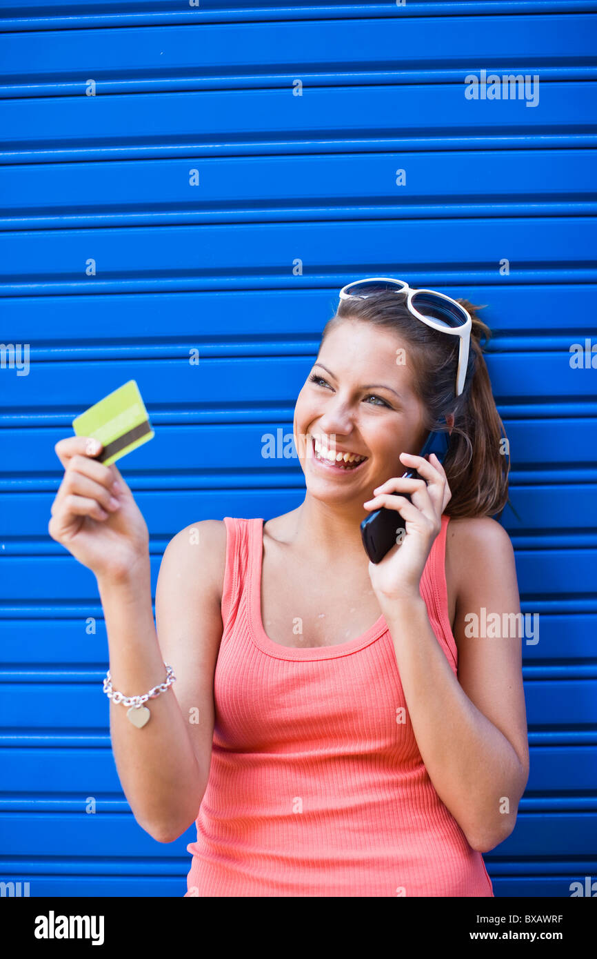 Young woman in front of blue wall, holding credit card and talking on phone Stock Photo