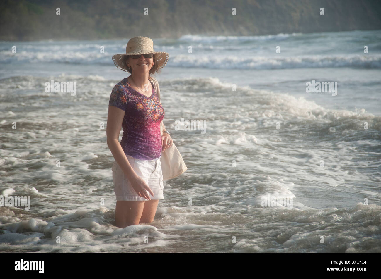 Woman in a hat smiling standing in the ocean surf at knee height Stock Photo