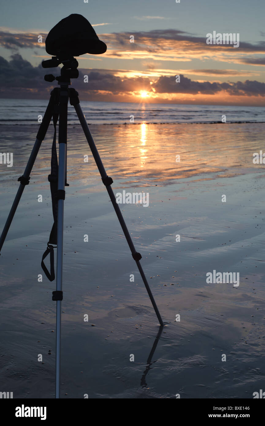 A photographers tripod stands on the wet beach in front of a sunset ...
