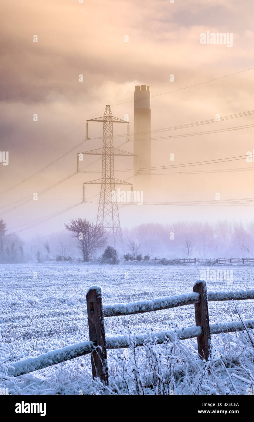 Power station chimney and electricity pylon showing through cold morning mist behind a frosty field Stock Photo