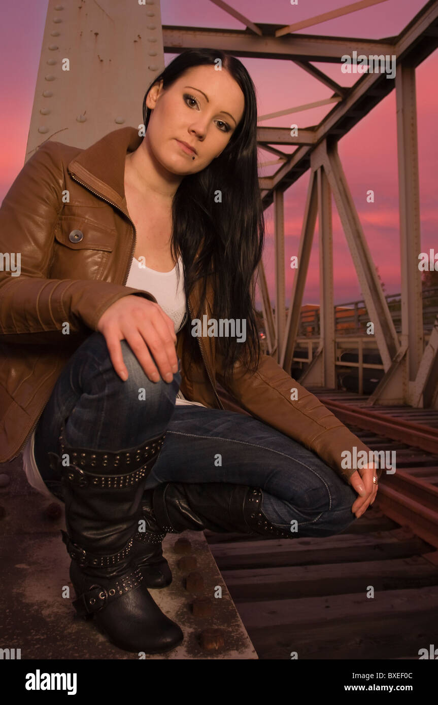 A long-haired young pretty woman in a leather jacket sits on a railroad bridge at sunset. Stock Photo