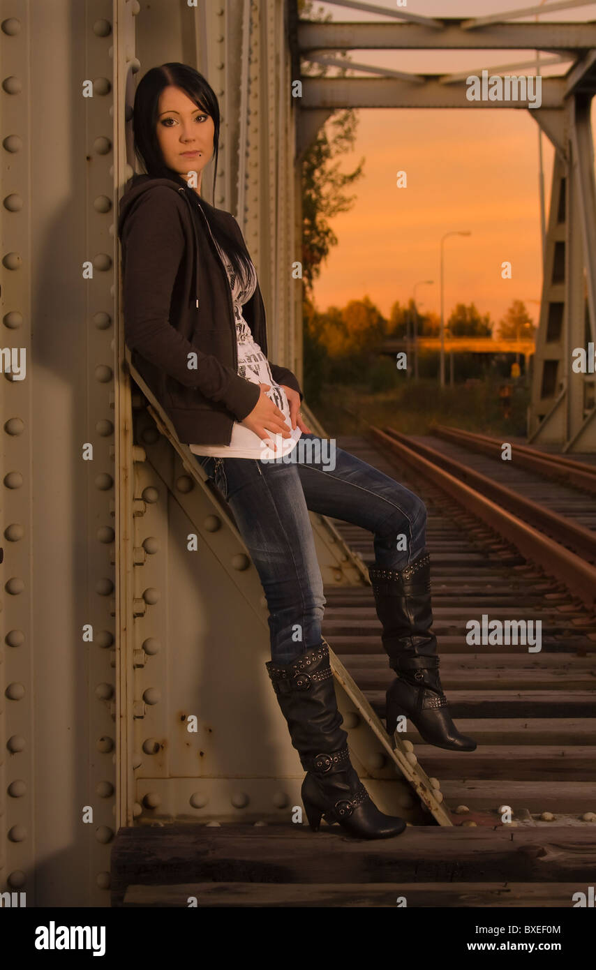A young woman  in jeans and boots and a leather jacket stands on a railroad bridge and leans against a beam. Stock Photo