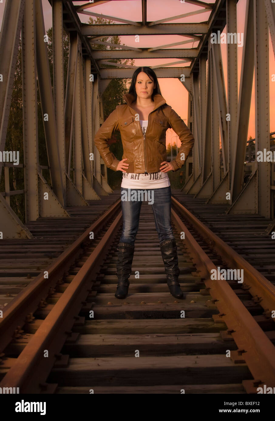 A young woman in a leather jacket standing on a railroad bridge in the evening sun. Stock Photo