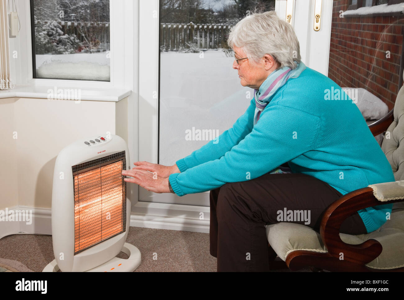 A senior woman pensioner OAP trying to keep warm warming hands indoors sat in front of a halogen electric fire with snow outside a window in winter UK Stock Photo