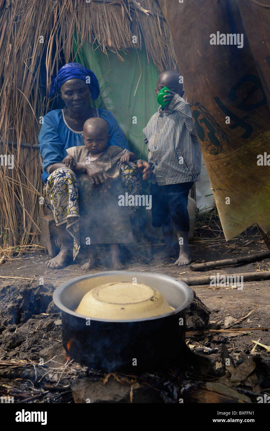 An internally displaced woman with her children heats food over bonfire in front of a makeshift straw hut in an IDP camp in North Kivu province DR Congo Africa Stock Photo