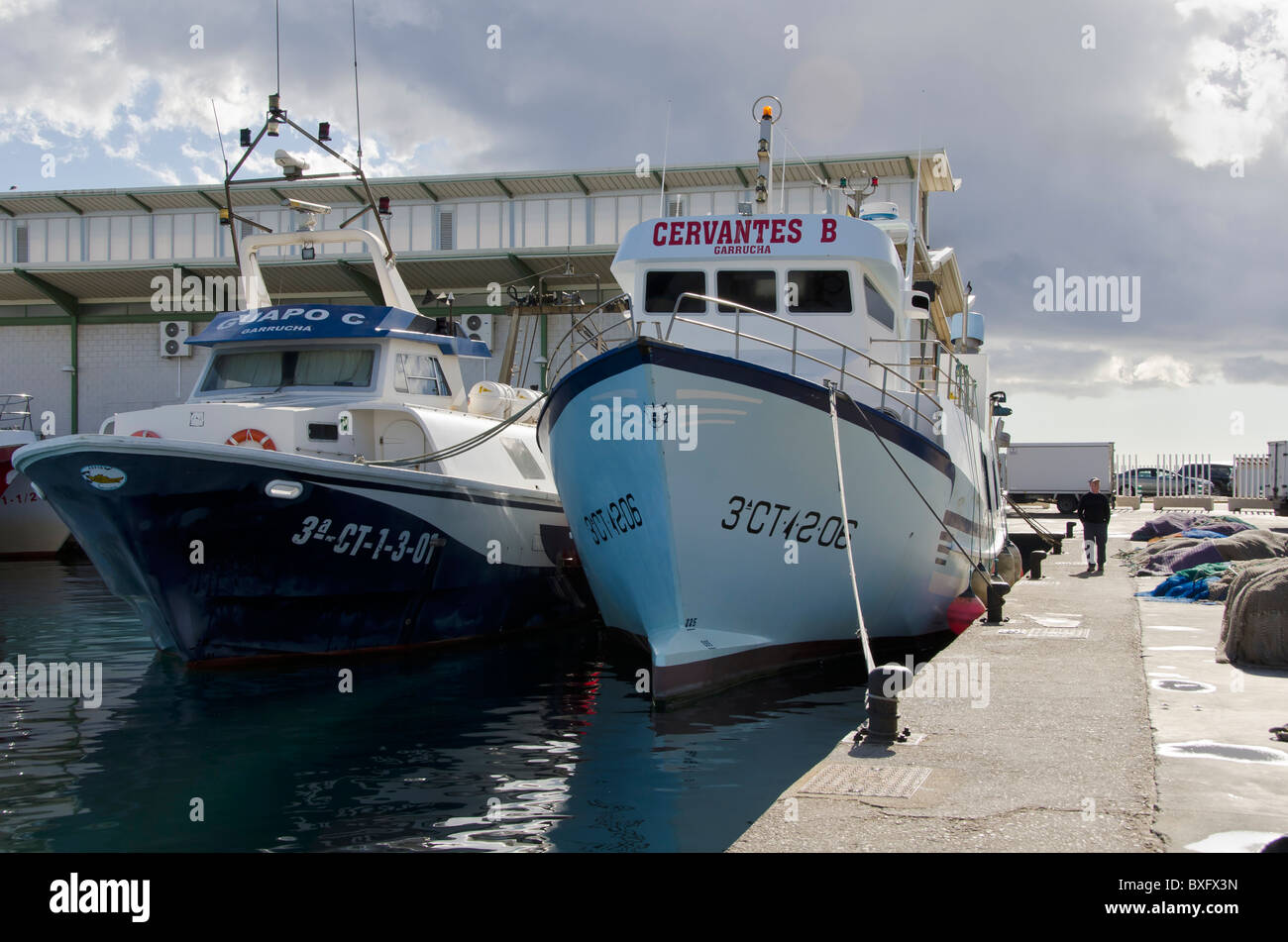Fishing Boats in Garrucha harbour, Spain on a Sunday lunchtime. Two boats are moored side by side in the harbour and a man is walking past. Stock Photo