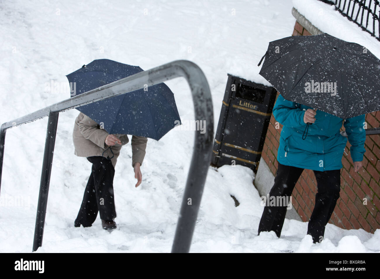 two women holding umbrellas walking up snow covered steps on a cold snowy winters day Belfast Northern Ireland Stock Photo