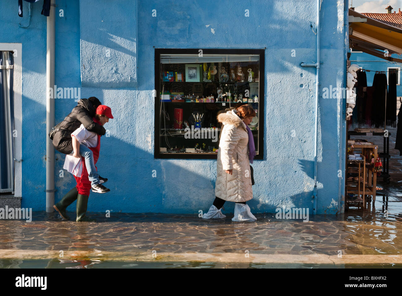 Woman carrying a friend while another walks with plastic bags on her feet on a street flooded by 'acqua alta', Burano, Italy, Eu Stock Photo
