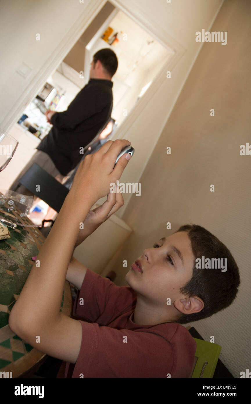 Ten year old boy sending a text message on his mobile phone while his father is busy in the kitchen. Stock Photo