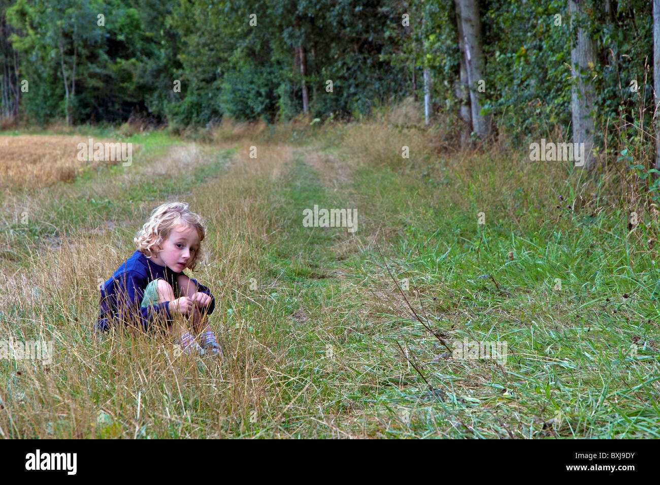 Four year old girl sitting alone in the grass beside a forest, France. Stock Photo