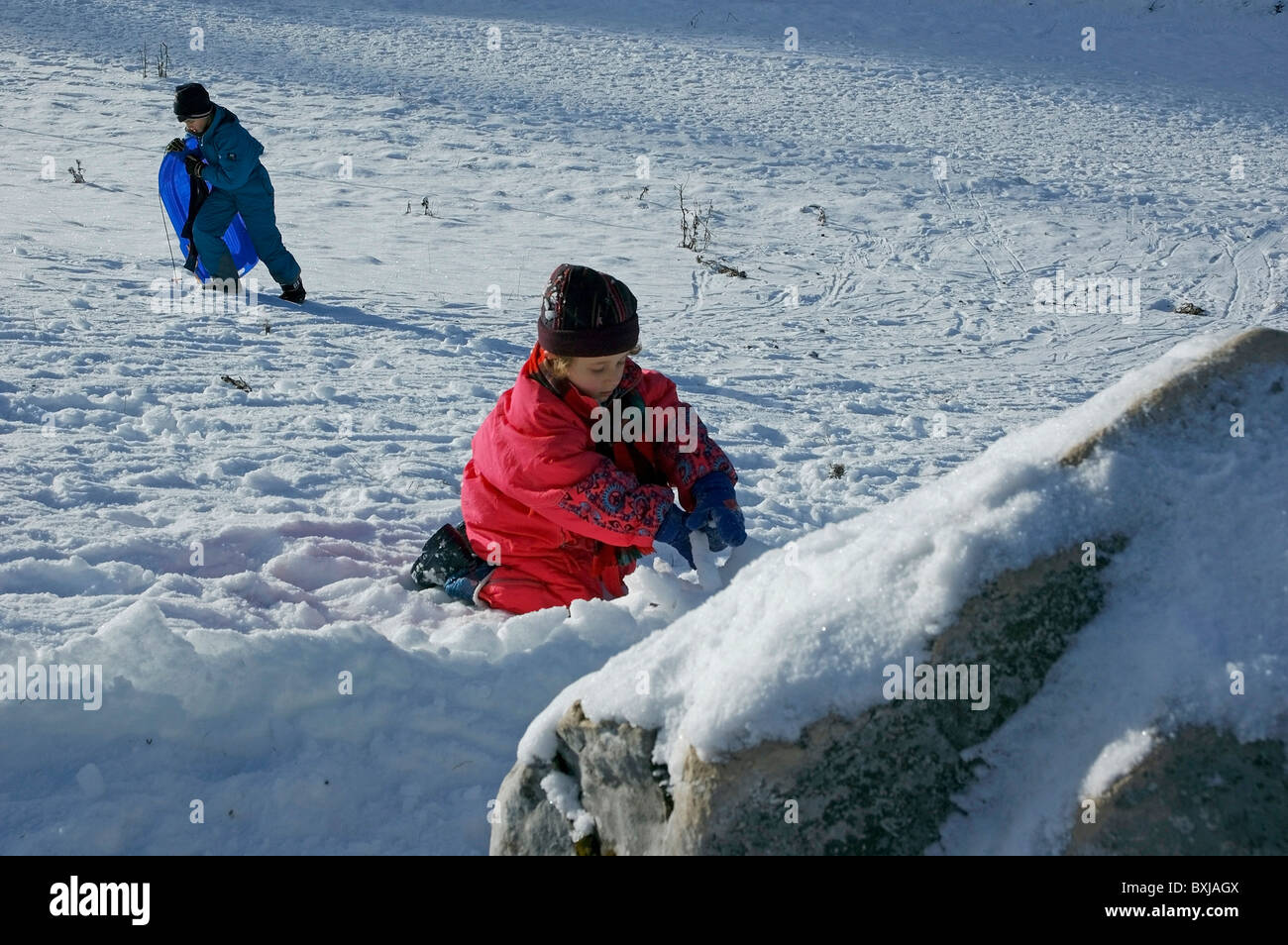 Little girl playing in snow by herself while her brother goes sledding. Stock Photo