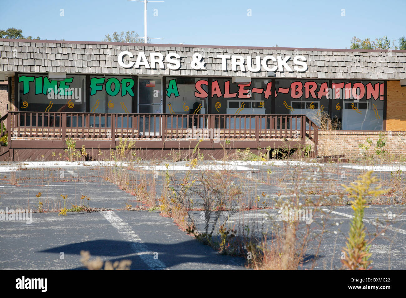 Empty abandoned car and truck dealer lot with weeds growing. Sign reads time for a sale a bration. Stock Photo