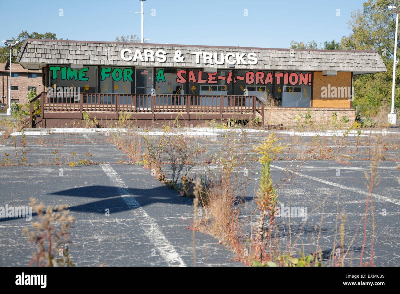 Empty abandoned car and truck dealer lot with weeds growing. Sign reads time for a sale a bration. Stock Photo