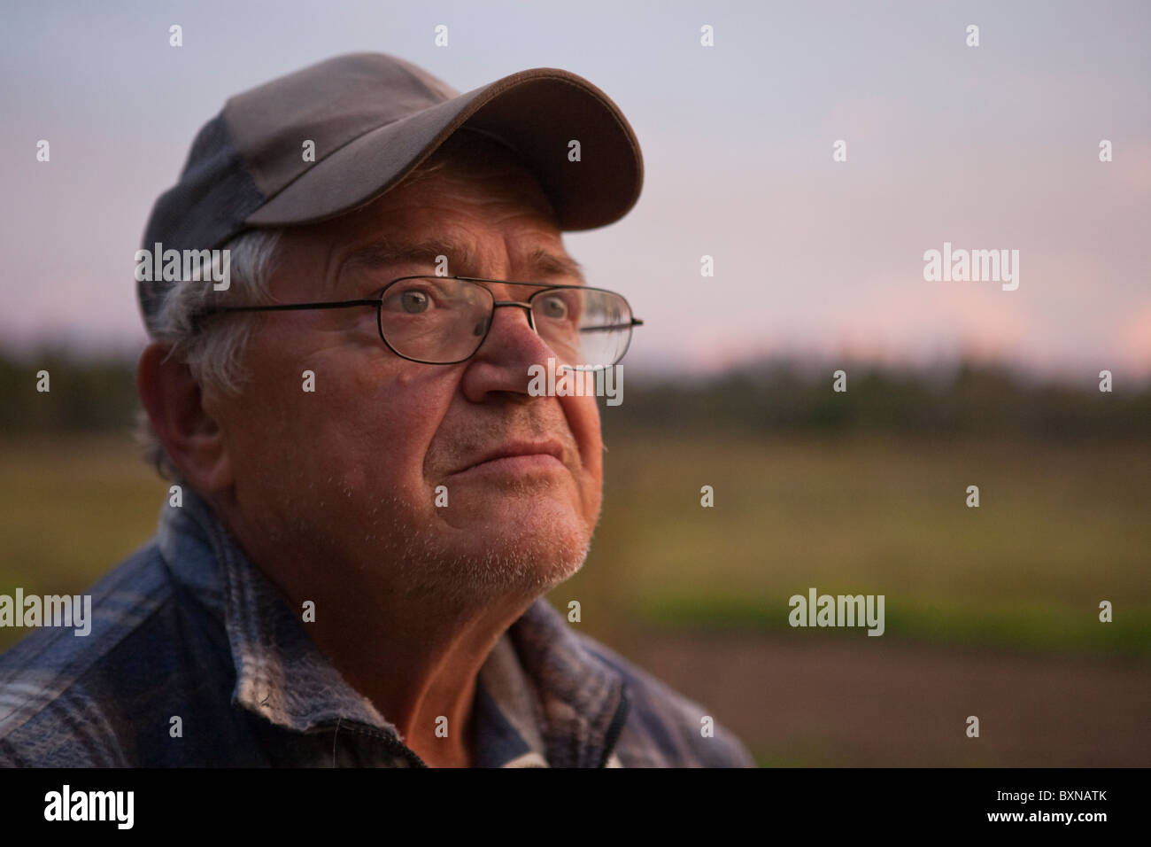 An elderly man with glasses and a cap stares pensively into the sky against the backdrop of the countryside Stock Photo