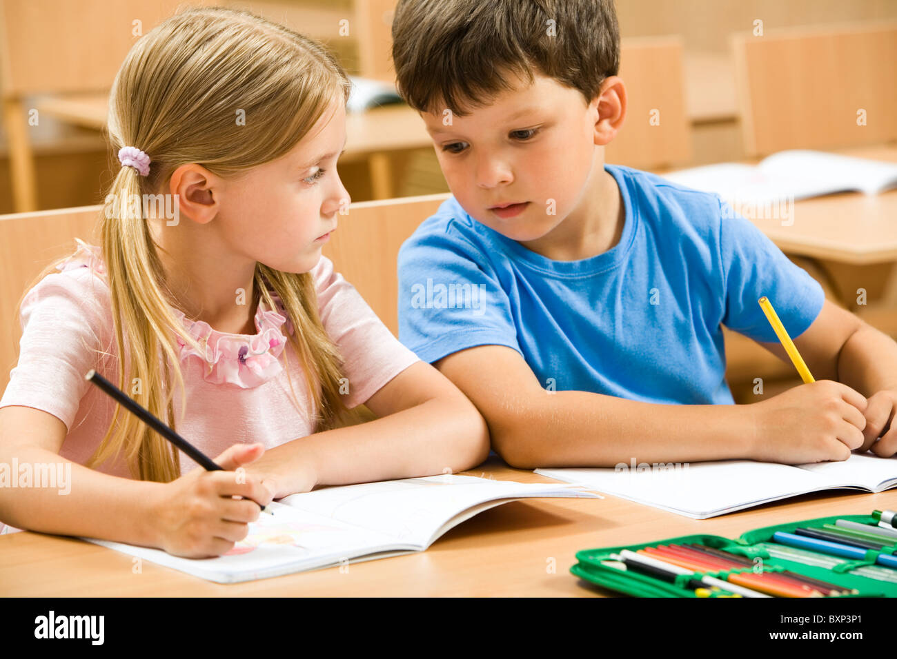 Portrait of two schoolchildren sitting by one desk during drawing lesson while boy looking at his mate’s copybook and girl watch Stock Photo