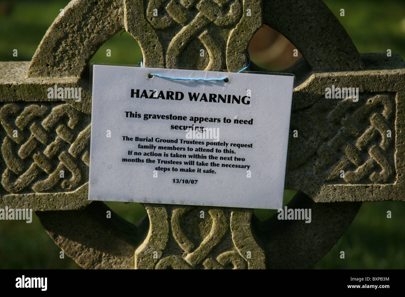 Hazard warning subsidence sign tied on a carved stone cross headstone in a graveyard Stock Photo
