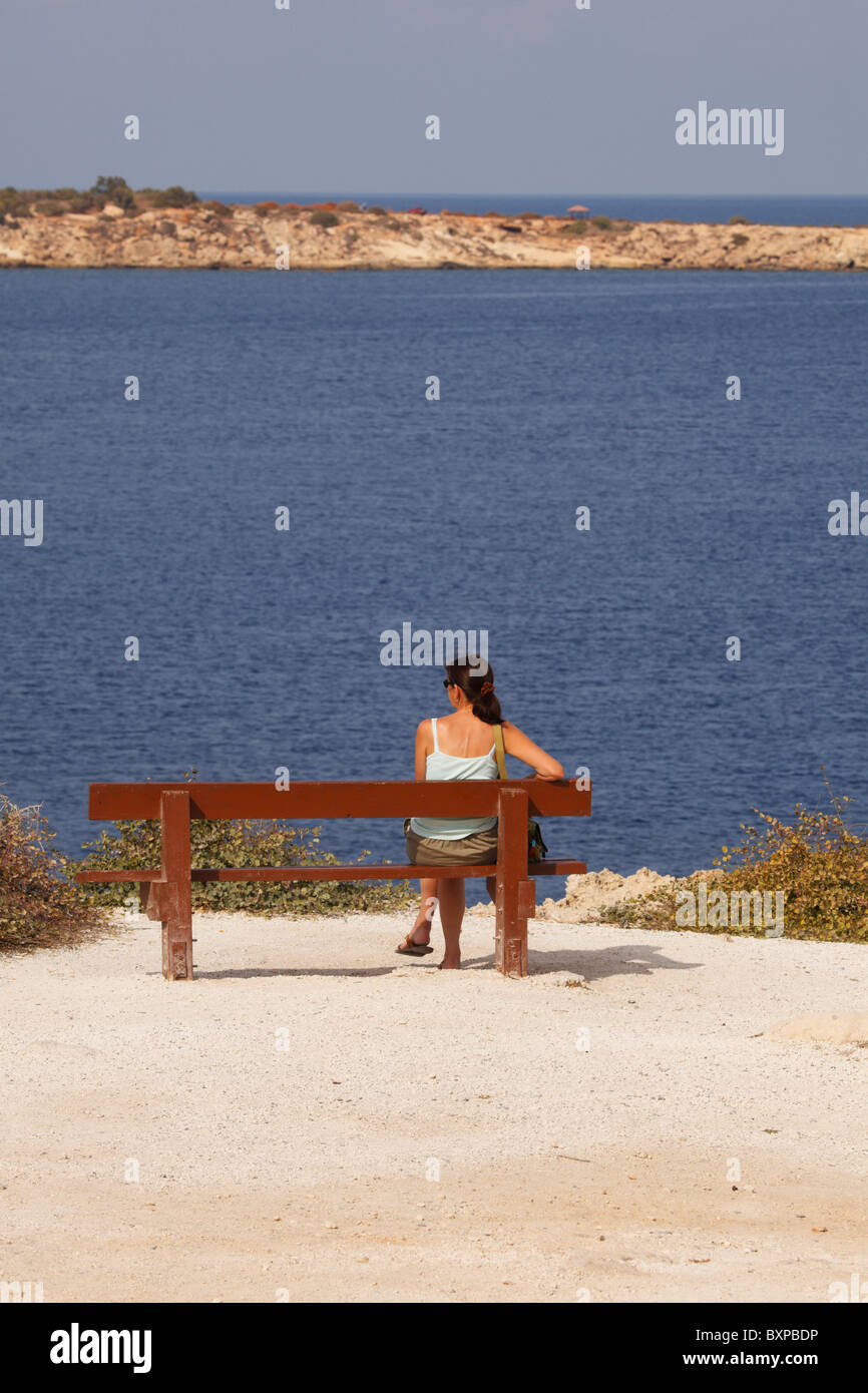 Female tourist sitting on a bench looking out to sea, Governors Beach, Cape Dolos, Cyprus Stock Photo