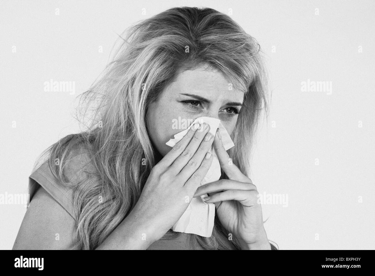 Young blond woman with a cold sneezing and blowing her nose on a tissue Stock Photo