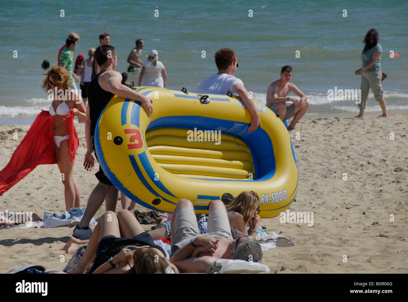 TWO YOUNG MALE CARRY RUBBER BOAT IN THE SEA SIDE RESORT DORSET IN ENGLAND Stock Photo