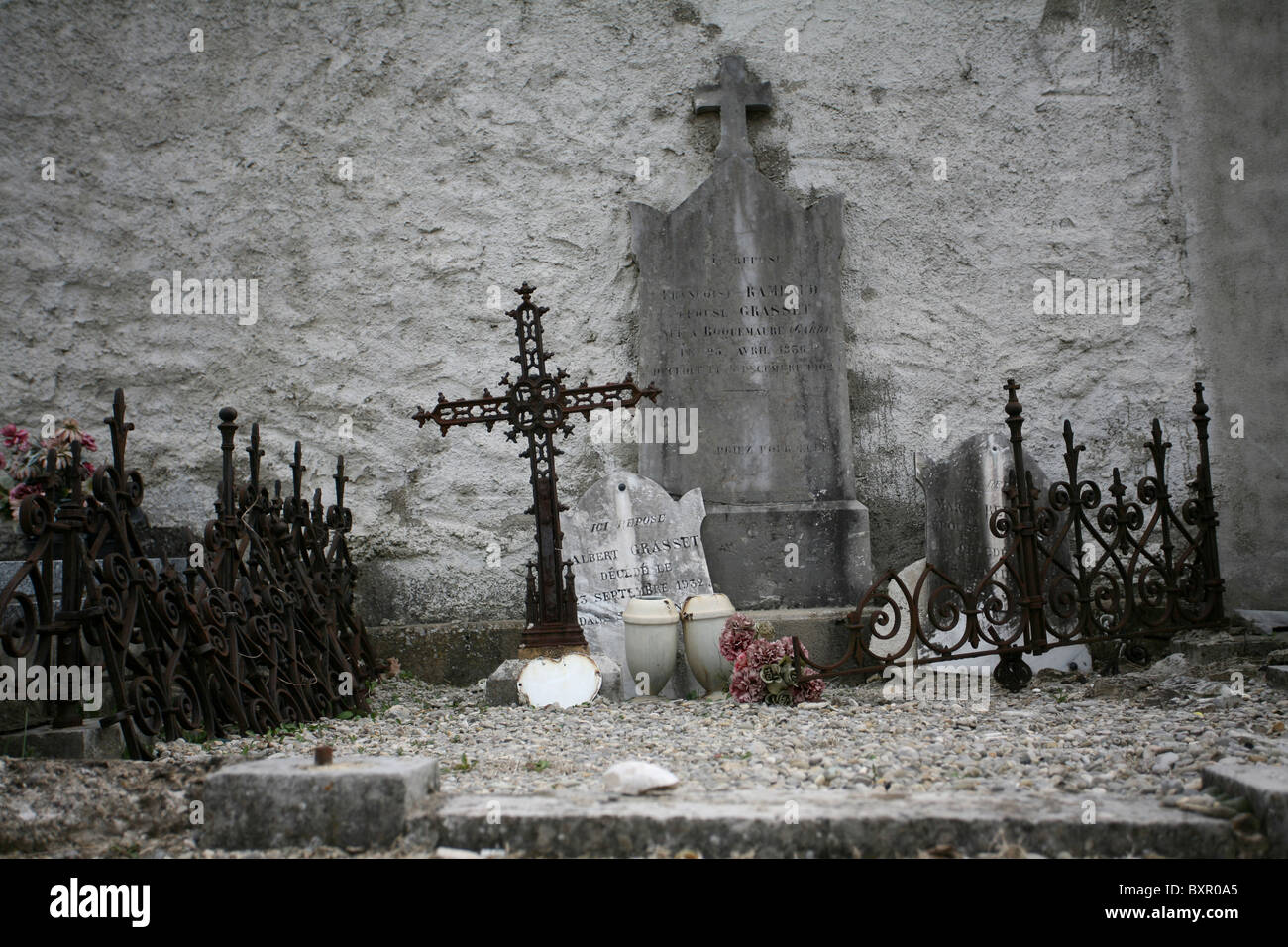 A gravesite with broken headstones, crosses, metal railings, and ornaments, against a wall in a French cemetery. Stock Photo