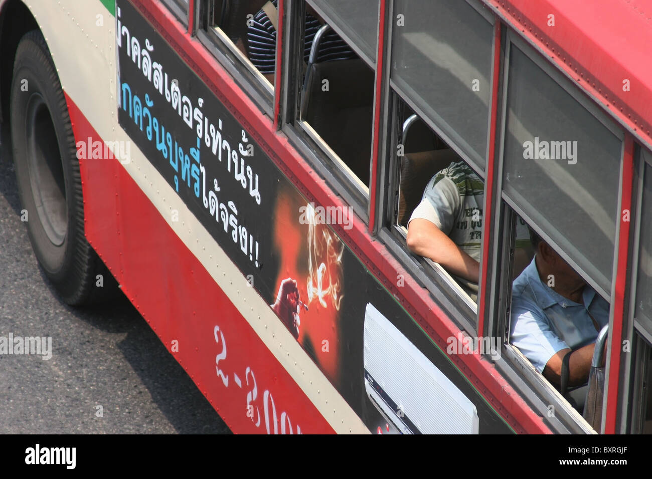 A bus with cigarette advertising is traveling with passengers sitting near open windows on a street in Bangkok, Thailand. Stock Photo